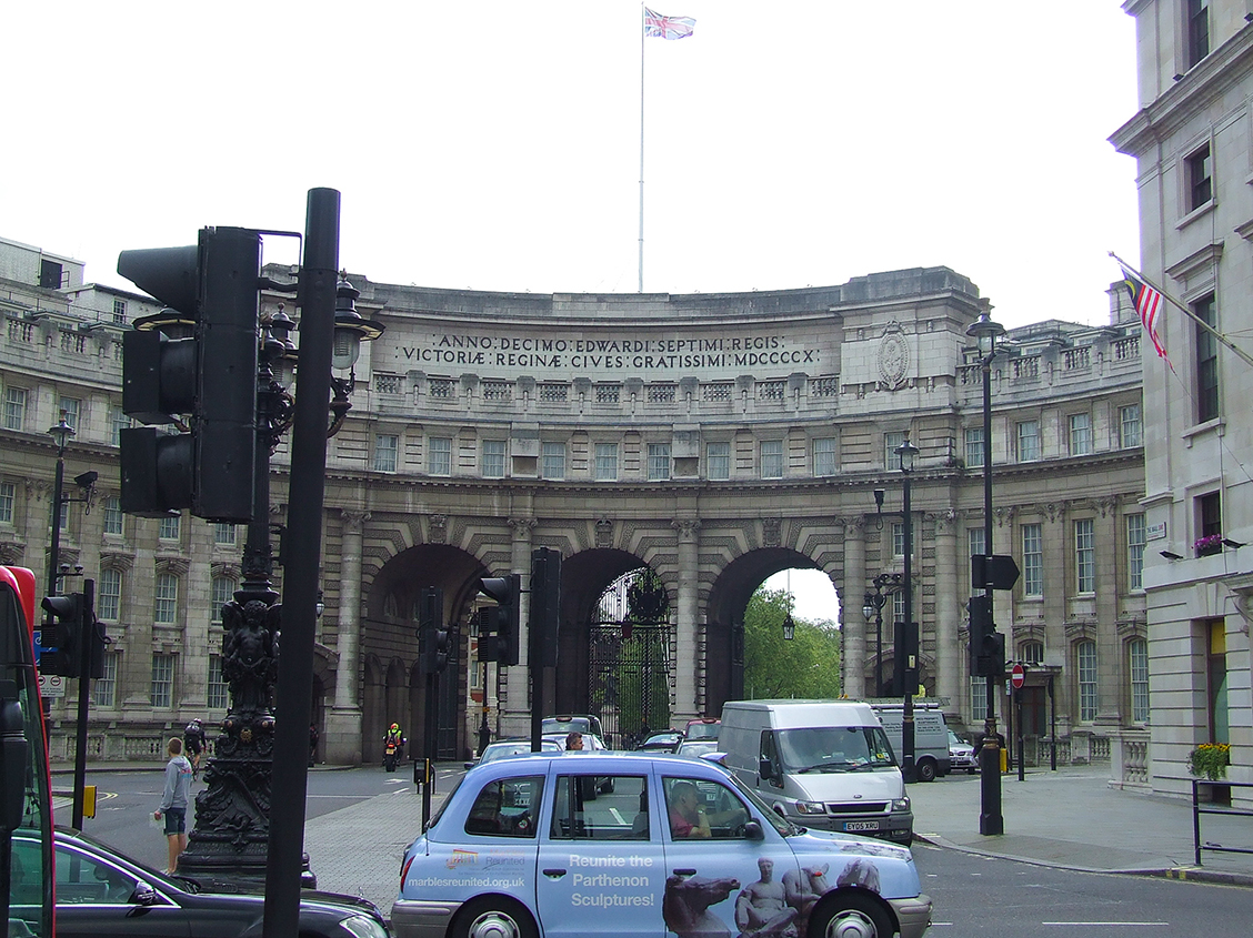 The image depicts the iconic Admiralty Arch in London, UK. The arch is a significant architectural landmark with a prominent inscription and serves as a gateway. The scene includes traffic, pedestrians, and buildings on either side. A notable detail is a taxi in the foreground with an advertisement for the reunification of the Parthenon Sculptures. The Union Jack and the American flag are visible, indicating the international context of the location.