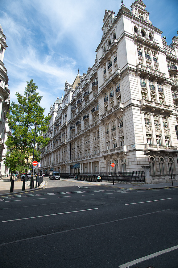 The image depicts a large, historic building with classical architecture, featuring tall columns and ornate details. The building is situated along a wide street with several vehicles parked in front, including a truck with the name 'Jones' on it. The sky is clear with a few clouds, and there are trees visible on the left side of the image..