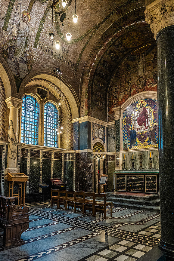 The image depicts an ornate interior of a historical building, likely a church or cathedral. The space features intricate mosaics, stained glass windows, and detailed marble work. The floor is adorned with a geometric tile pattern, and the room is furnished with wooden pews and a pulpit. The overall atmosphere is one of grandeur and reverence, highlighted by the elaborate decorations and architectural details.