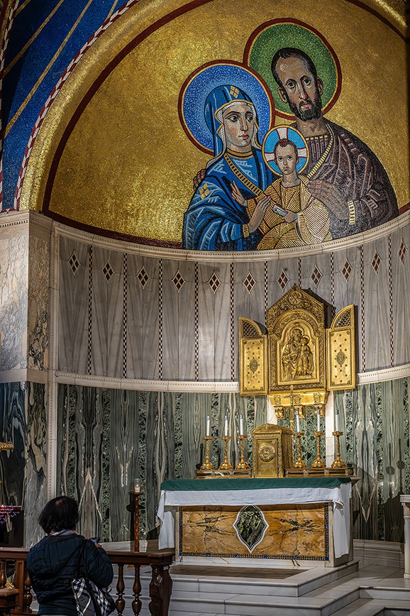 The image depicts an interior of a church or chapel, featuring a person kneeling in prayer. The focal point is a beautifully adorned altar with religious icons and artifacts. Above the altar, there is a large mosaic artwork depicting religious figures, likely the Holy Family, with intricate details and vibrant colors. The setting is richly decorated with gold and other ornate elements, creating a serene and reverent atmosphere.