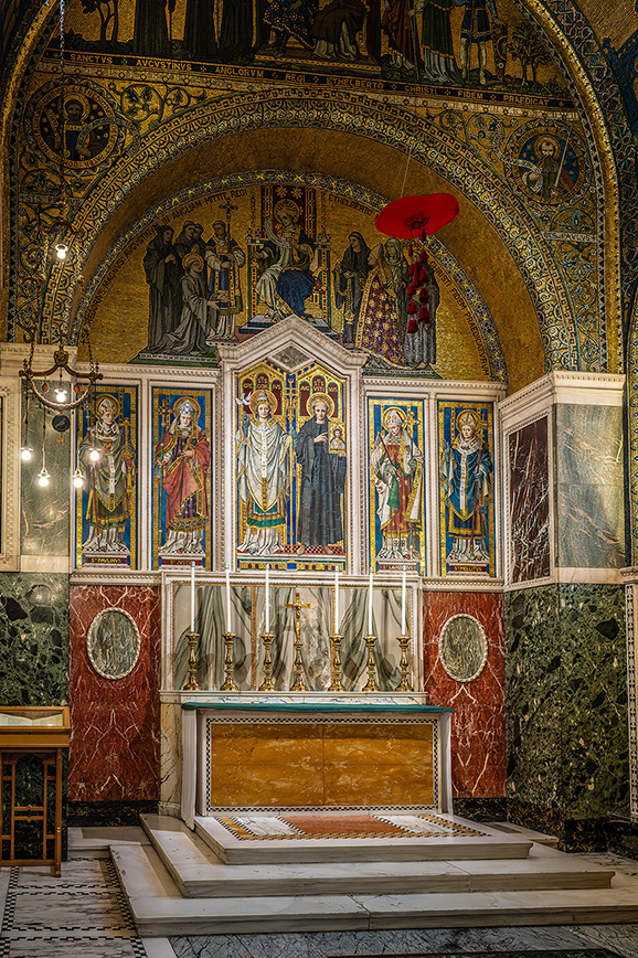 The image depicts an ornate altar within a church, featuring intricate mosaics and detailed artwork. The altar is adorned with religious figures and surrounded by elaborate decorations, including a prominent red umbrella-like structure above the central figure. The setting is rich in historical and artistic elements, reflecting a significant religious and cultural heritage.