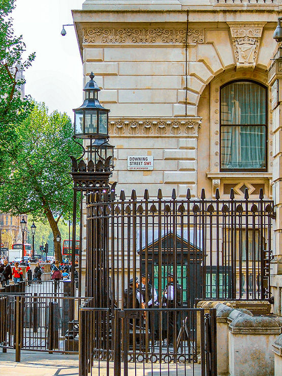 The image depicts the entrance to Downing Street in London, UK. The entrance features a black wrought iron gate with a guard post, and a traditional black lantern. The building has a classical architectural style with stone walls and large windows. There are people and police officers visible near the entrance, indicating security presence. The street sign 'Downing Street SW1' is prominently displayed on the building.
