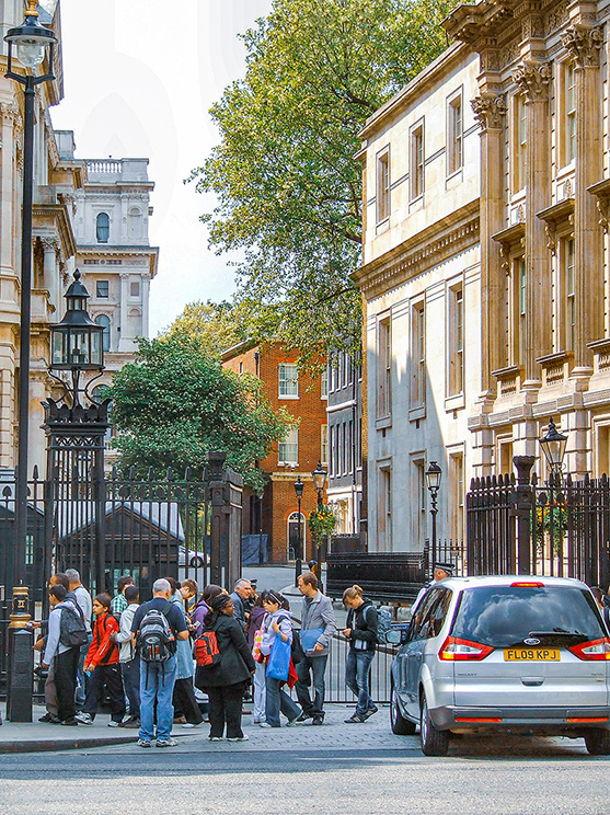 he image depicts a busy street scene in an urban area with historic architecture. People are gathered near a gated entrance, possibly tourists or visitors. The street is lined with tall, ornate buildings and trees. A car is parked on the right side of the street. The overall atmosphere suggests a mix of historical significance and modern activity.