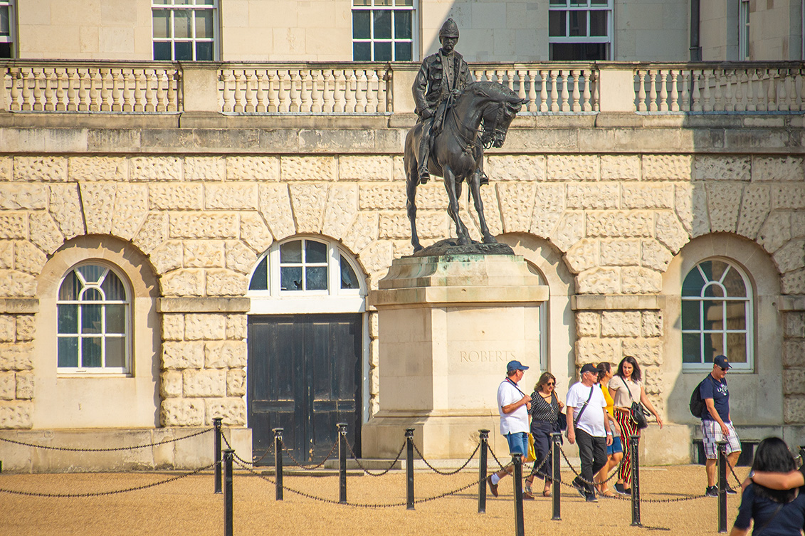 The image depicts a statue of a man on horseback in front of a historic building. The statue is mounted on a pedestal with the name 'ROBERT' inscribed on it. Several people are seen walking and standing around the statue, and the building features large arched windows and a prominent entrance door. The setting appears to be a public or tourist area, possibly a museum or a historic site.