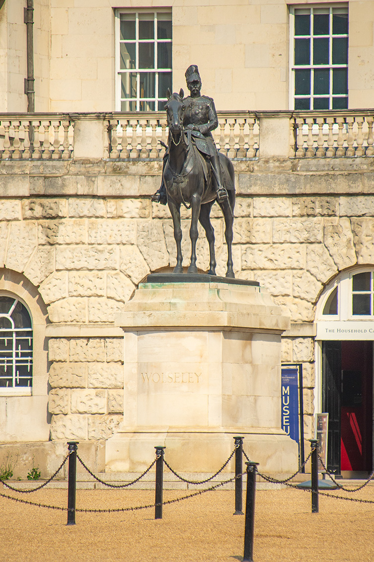The image shows a statue of a man on horseback in front of a building. The statue is mounted on a stone pedestal with the name 'Wolseley' inscribed on it. The building behind the statue has a sign that reads 'The Household Cavalry Museum'. The statue and the building are surrounded by a cordoned-off area with chains supported by black posts.