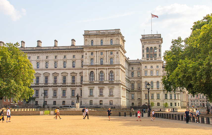 The image depicts a large, historic building with classical architecture, featuring multiple windows and ornate details. The building is surrounded by a spacious, open area where people are walking and engaging in various activities. The scene is set on a clear day with a blue sky and some clouds. Trees are visible on the sides, adding greenery to the urban setting. A flag is flying atop the building, indicating its significance.