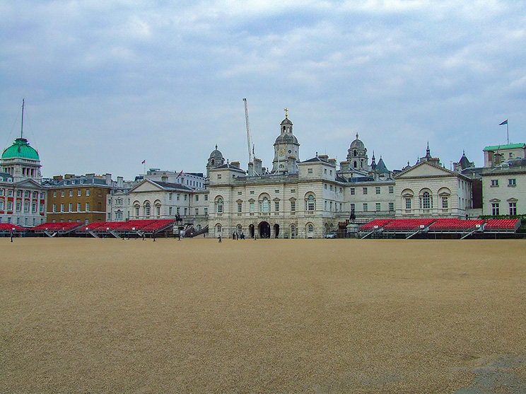 The image depicts Horse Guards in London, a historic building known for its architectural significance and role in British military history. The building features a central archway and is flanked by a large, open parade ground. The structure is characterized by its classical design, including domed roofs and symmetrical facades. Horse Guards serves as a significant landmark and tourist attraction in the city.