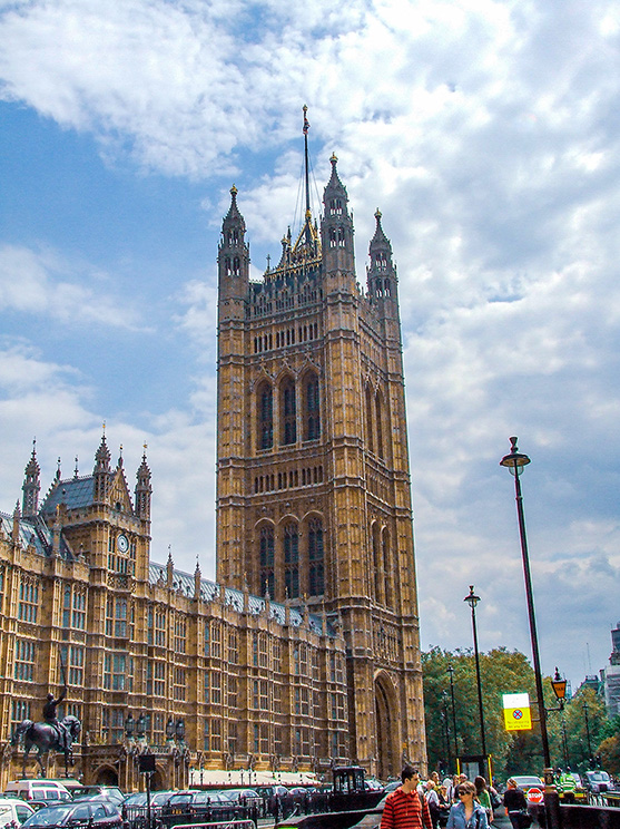 The image depicts the Palace of Westminster, a renowned landmark in London, United Kingdom. The photograph showcases the intricate Gothic architecture of the building, including the iconic Victoria Tower. The scene is bustling with activity, featuring numerous people walking on the street and several vehicles, including cars and buses, parked along the road. The sky is partly cloudy, adding to the overall atmosphere of the image. The photograph captures the grandeur and historical significance of this famous political and architectural site.