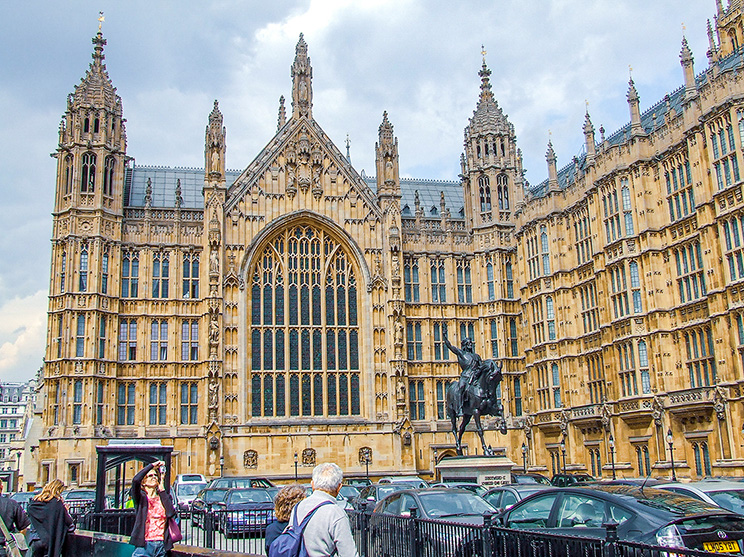 The image depicts the Palace of Westminster in London, UK, featuring its iconic Gothic architecture with detailed stone carvings and large pointed arches. In the foreground, there is a statue of a prominent figure on horseback, surrounded by parked cars and several people walking and taking photos.