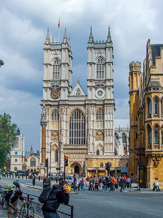 The image depicts Westminster Abbey, a large, mainly Gothic abbey church in London, England. It is one of the most notable religious buildings in the UK and has been the traditional place of coronation and burial site for English and, later, British monarchs. The image shows the intricate architectural details of the abbey, including its twin towers, large windows, and clock face. There are people and vehicles around the abbey, indicating it is a busy and popular whitehall.