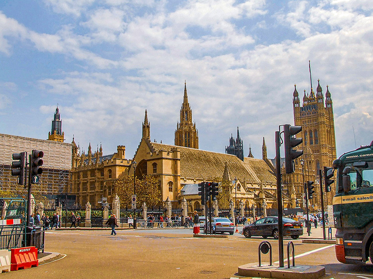 The image depicts a busy street scene in front of a large, historic building with Gothic architecture, likely a cathedral or significant landmark. The building features tall spires, intricate stonework, and large windows. There are several people walking and a few vehicles, including a bus and cars, on the street. Traffic lights and street signs are visible, indicating a well-regulated urban area. The sky is clear with some clouds, suggesting a pleasant day.