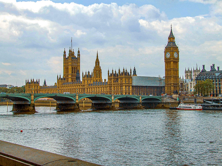 The image depicts the iconic Palace of Westminster and the Elizabeth Tower, commonly known as Big Ben, in London. The scene includes the Westminster Bridge spanning the River Thames, with a boat navigating the river. The sky is partly cloudy, and the architectural details of the Gothic Revival style buildings are prominently displayed.