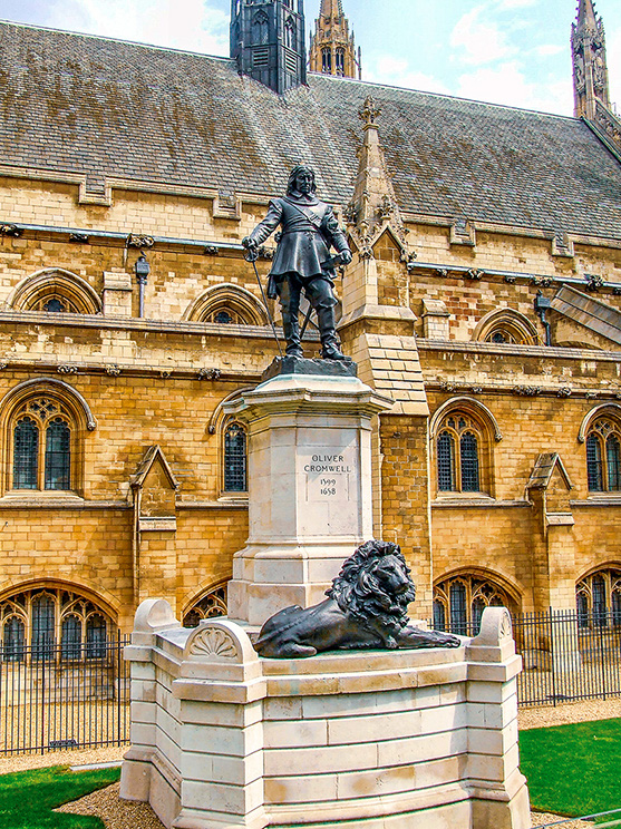 The image depicts a statue of Oliver Cromwell, a historical figure, situated in front of a large, ornate building with Gothic architecture. The statue stands on a pedestal with a lion at its base. The pedestal bears the inscription 'Oliver Cromwell 1599-1658'. The building behind the statue features pointed arches, intricate stonework, and tall, narrow windows, indicative of Gothic style.
