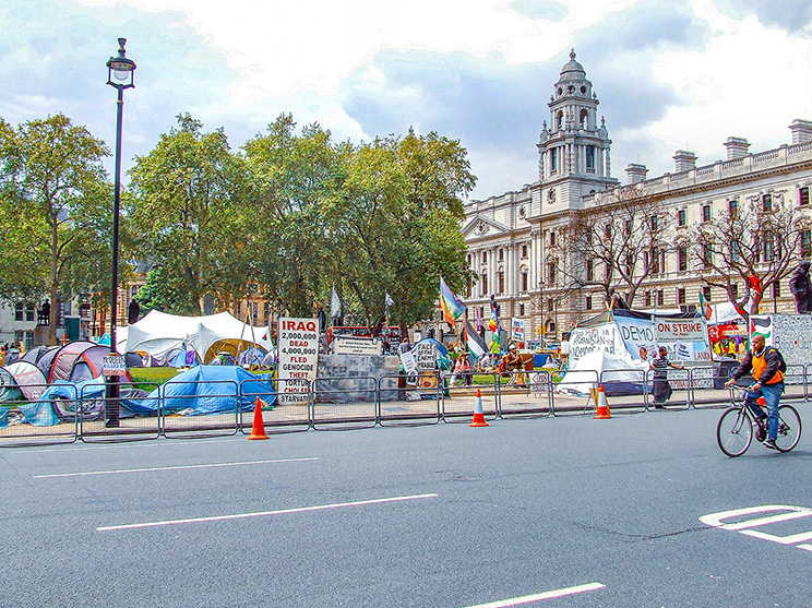 The image depicts a protest or demonstration taking place in front of a large, historic building. Various tents and signs are set up, with one prominent sign mentioning 'IRAQ.' There are barriers and traffic cones separating the protest area from the street. A cyclist is seen riding past the protest on the street. The scene is set on a wide road with trees and a lamppost visible in the background.