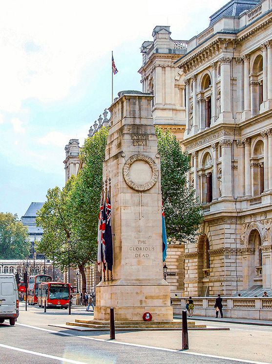The image depicts a war memorial monument situated in a busy urban area, likely in London. The monument is adorned with flags and features an inscription that reads 'The Glorious Dead.' The surrounding architecture is grand and classical, with a prominent building in the background. The street is lined with trees and bustling with vehicles, including a red double-decker bus and a white van.