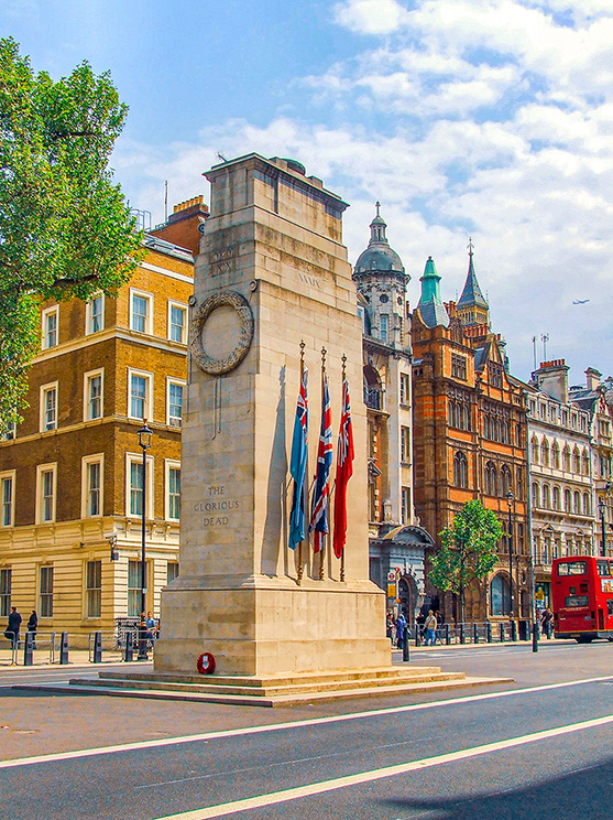 The image depicts a war memorial in a bustling city square, featuring a tall stone monument with a circular design at the top and several flags, including the Union Jack. The monument is inscribed with 'The Glorious Dead.' The background showcases historic buildings with classic European architecture, including domes and spires. A red double-decker bus and pedestrians are also visible, indicating a lively urban environment.