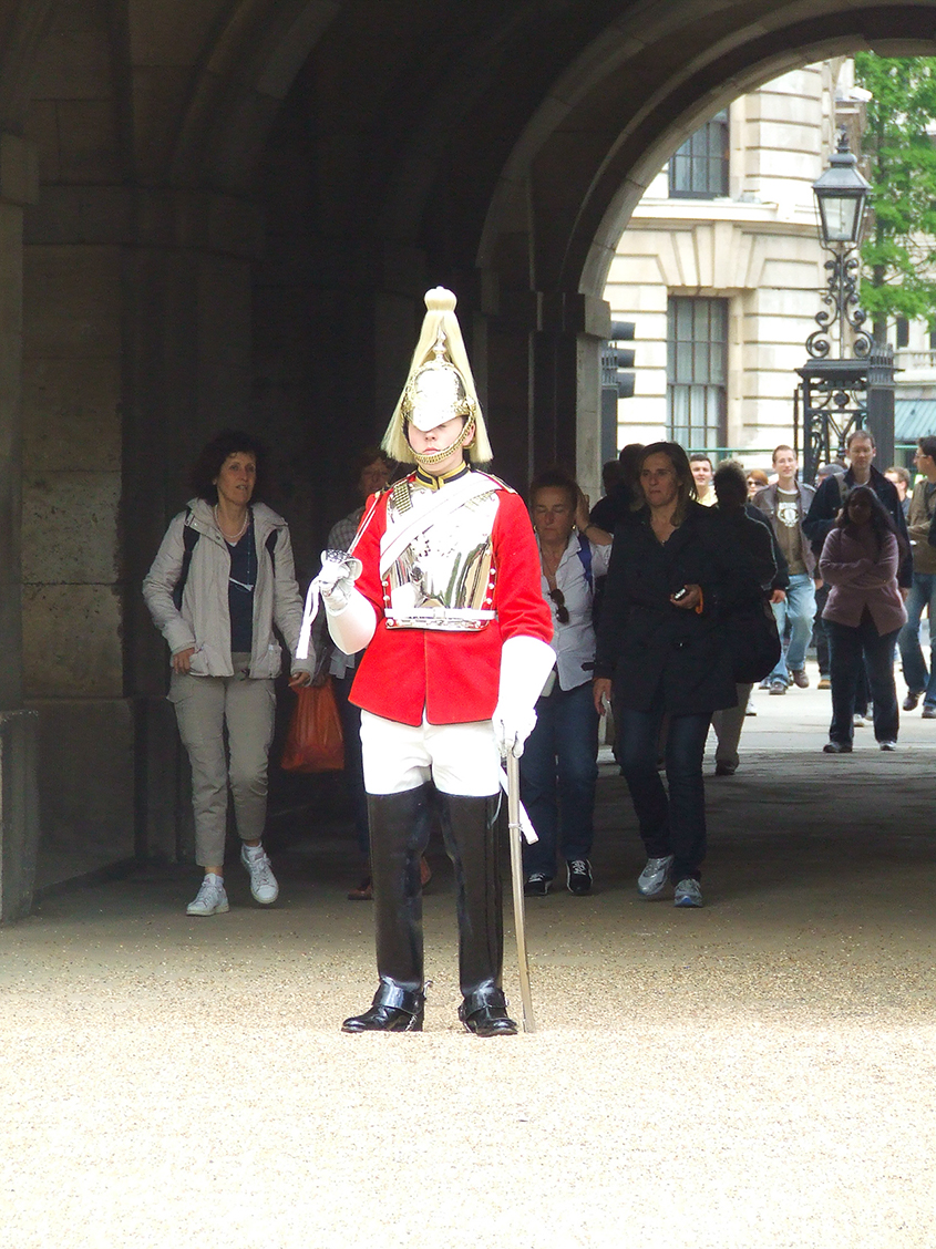 The image depicts a ceremonial guard, likely a member of the Queen's Guard, standing at attention in a traditional red uniform with white accents and a tall bearskin hat. The guard is holding a weapon and white glove, positioned in front of an archway. Several people are observing the guard, suggesting a tourist setting, possibly at a historic or royal site in the UK.