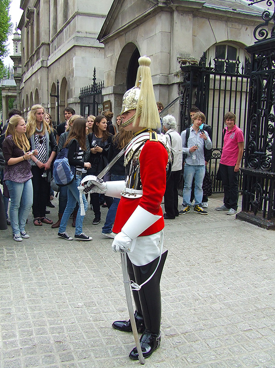 The image depicts a ceremonial guard dressed in a traditional red and white uniform with a tall bearskin hat, standing at attention in front of a historic building with a crowd of tourists observing and taking pictures.