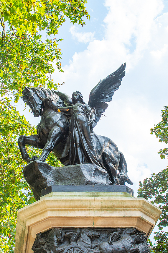 The image depicts a statue of a winged figure riding a horse. The statue is mounted on a stone pedestal, surrounded by trees with green leaves and a partly cloudy sky in the background.