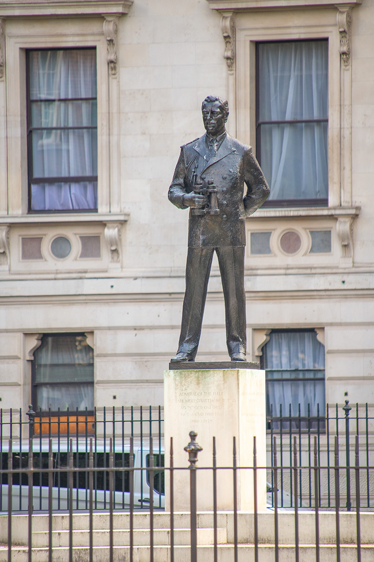 The image depicts a statue of a man standing in front of a building. The statue is mounted on a pedestal with an inscription. The man in the statue is holding an object, possibly binoculars or a small case, and is dressed in 19th-century attire. The building behind the statue has large windows with curtains and decorative elements around the windows. The statue is enclosed within a black metal fence.