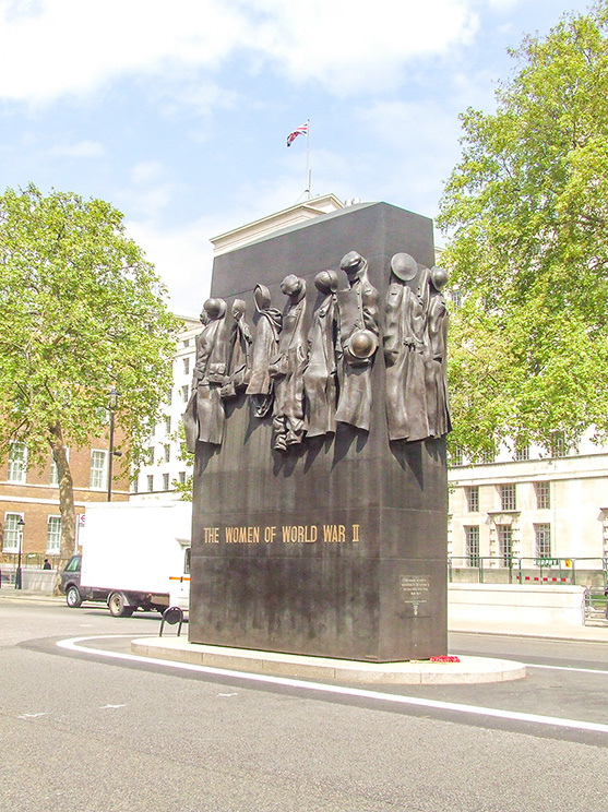 The image depicts a monument dedicated to the women of World War II. The monument features a large, dark stone structure with relief sculptures of women in various roles and attire, symbolizing their contributions during the war. The inscription on the monument reads 'The Women of World War II.' The setting appears to be an urban area with trees and buildings in the background, and a flag flying atop the monument.