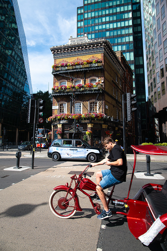 The image depicts an urban scene with a person riding a red tricycle on a city street. The street is lined with modern high-rise buildings, and there is an older, ornate building adorned with flower boxes in the background. The scene includes a taxi and several pedestrians, indicating a bustling city environment.