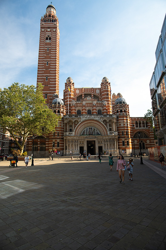 The image depicts a large, ornate building with a tall tower, likely a cathedral or church, characterized by its red brick and stone construction. The structure features intricate architectural details, including arches, domes, and decorative elements. The scene is set in a spacious plaza with a few people walking around, indicating it is a public and possibly tourist-friendly area. The sky is clear, suggesting a sunny day.
