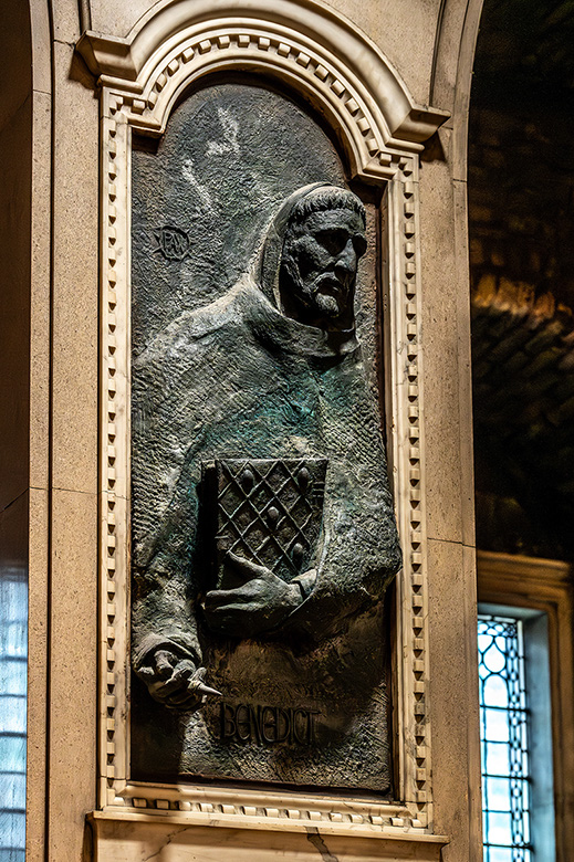 The image depicts a bronze relief sculpture of a robed figure, likely a historical or religious figure, set within an ornate stone frame. The figure is holding an object that appears to be a book or a shield with a cross, suggesting a connection to Christianity. The name 'Benedict' is inscribed at the bottom, indicating that the figure is St. Benedict. The sculpture is located in an interior space with stained glass windows, possibly a church or a monastery.