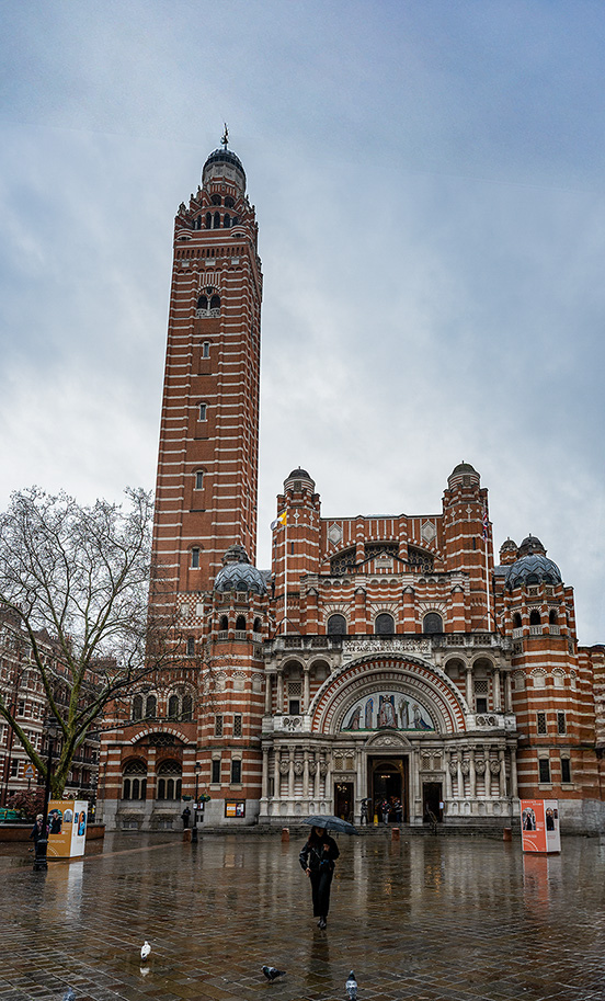 The image depicts a large, ornate building with a tall tower, likely a cathedral or church, characterized by its red brick and stone construction. The architecture features intricate details, arched windows, and decorative elements. The scene is set on a rainy day, with a person holding an umbrella in the foreground, and the ground appears wet and reflective. The sky is overcast, adding to the gloomy atmosphere. The building's design suggests it is of significant historical and architectural importance.