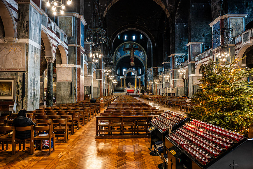 The image depicts the interior of a grand, ornate church or cathedral. The space is characterized by high ceilings, large arches, and intricate decorations. Rows of wooden pews are arranged in the main aisle, leading to an altar adorned with a large crucifix. The church is decorated with a Christmas tree and festive lights, indicating a holiday season setting. The overall atmosphere is solemn and reverent, with a few individuals present, suggesting a quiet, reflective moment.