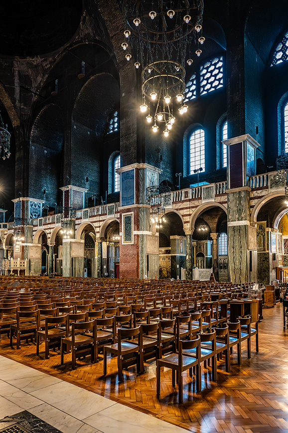 The image depicts the interior of a grand, empty church or cathedral. The space is characterized by high, arched ceilings, large chandeliers, and rows of wooden pews. The architecture features intricate stonework, arches, and large stained-glass windows that allow natural light to filter into the space. The overall atmosphere is serene and majestic, highlighting the historical and architectural significance of the building.