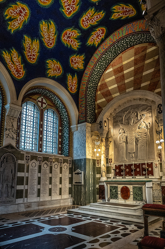 The image depicts an ornate interior of a church or cathedral. The ceiling features colorful decorations with names inscribed on flames, possibly commemorating individuals. The walls are adorned with intricate patterns and stained glass windows. At the center, there is an altar with detailed carvings and a memorial plaque, suggesting it is a place of worship and remembrance.