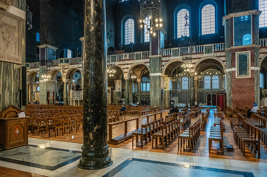 The image depicts the interior of a grand, ornate church or cathedral. The space is characterized by high ceilings, large stained-glass windows, and marble columns. Rows of wooden pews are arranged neatly, leading up to an altar area. The architecture features intricate details, including arches, chandeliers, and decorative elements. The overall atmosphere is serene and majestic.