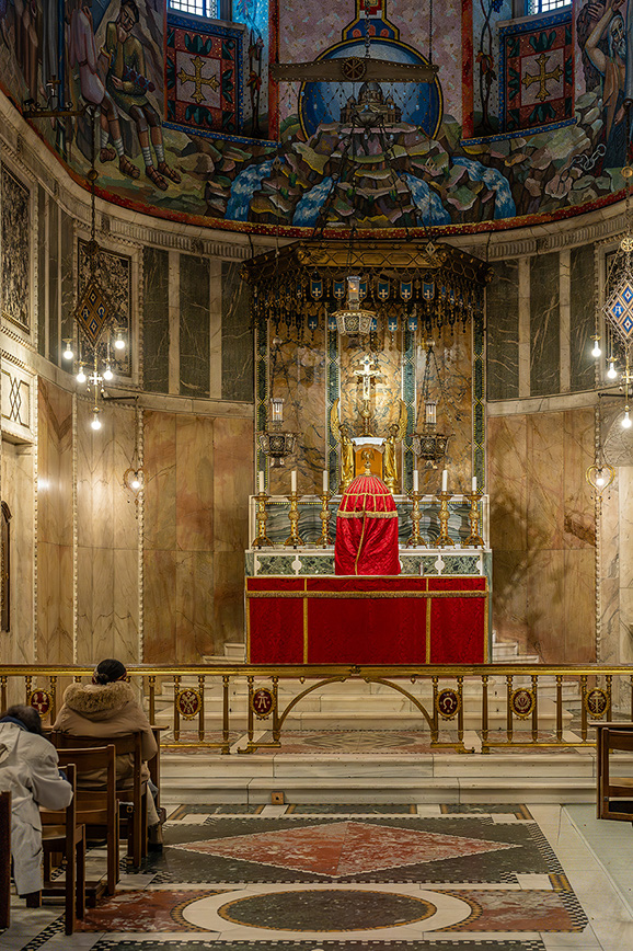 The image depicts an ornate, circular church interior with intricate mosaics and frescoes adorning the ceiling and walls. The focal point is an elevated altar draped in red, with a statue of a religious figure above it. Two individuals are seated in the foreground, facing the altar, suggesting a moment of prayer or reflection. The floor features a detailed, circular mosaic design, and the overall atmosphere is serene and reverent.