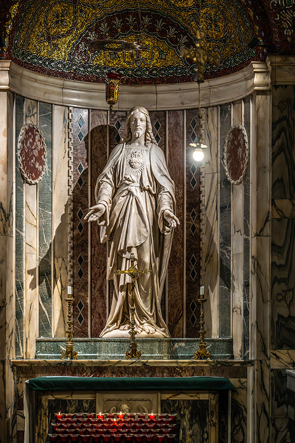 The image depicts a statue of Jesus Christ inside a church, adorned with intricate decorations and surrounded by candles.