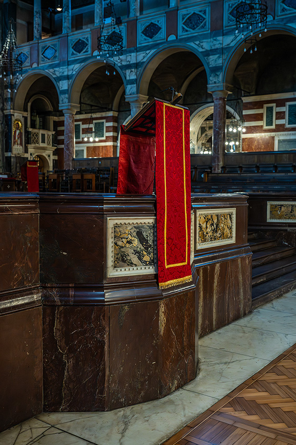The image depicts an interior view of a church or cathedral. The focal point is a wooden pulpit adorned with a red cloth and gold trim. The pulpit is decorated with intricate artwork and is situated in a large, ornately designed space featuring arches, marble floors, and detailed architectural elements. The setting suggests a place of worship with historical and artistic significance.