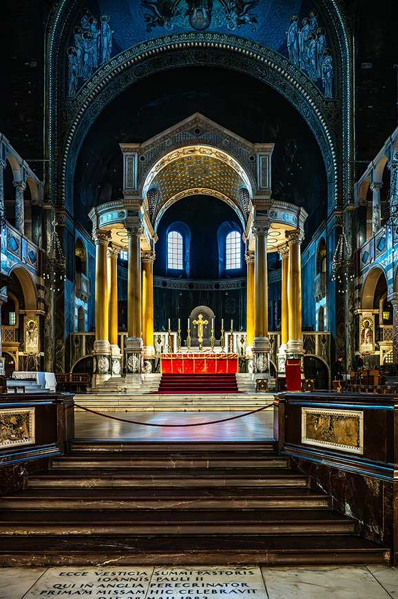 The image depicts the interior of a richly decorated church, focusing on the altar area. The altar is adorned with a red cloth and surrounded by golden columns and intricate mosaics. The architecture features arches and detailed artwork on the walls and ceiling. The overall atmosphere is solemn and reverent, typical of a place of worship.