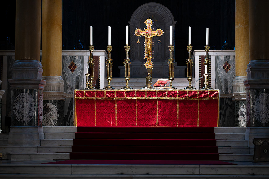 The image depicts an altar inside a church, adorned with a red and gold cloth, several lit candles, and a prominent crucifix at the center. The altar is elevated with steps and flanked by ornate columns.