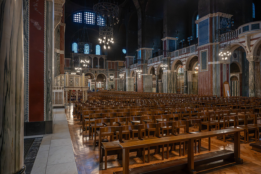 The image depicts the interior of a grand, ornate church or cathedral. The space is characterized by high ceilings, intricate architectural details, and rows of wooden pews. The lighting is dim, with chandeliers providing a warm glow. The walls and columns are adorned with detailed patterns and designs, and there are large, stained-glass windows that allow natural light to filter into the space. The overall atmosphere is serene and reverent.