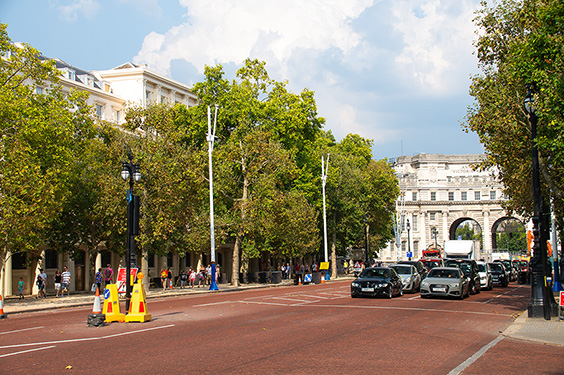 The image depicts a wide, tree-lined street with a mix of pedestrians and vehicles. The street is bordered by historic buildings, and there is a notable archway structure in the background. The scene suggests a well-maintained urban area with a blend of architectural styles.