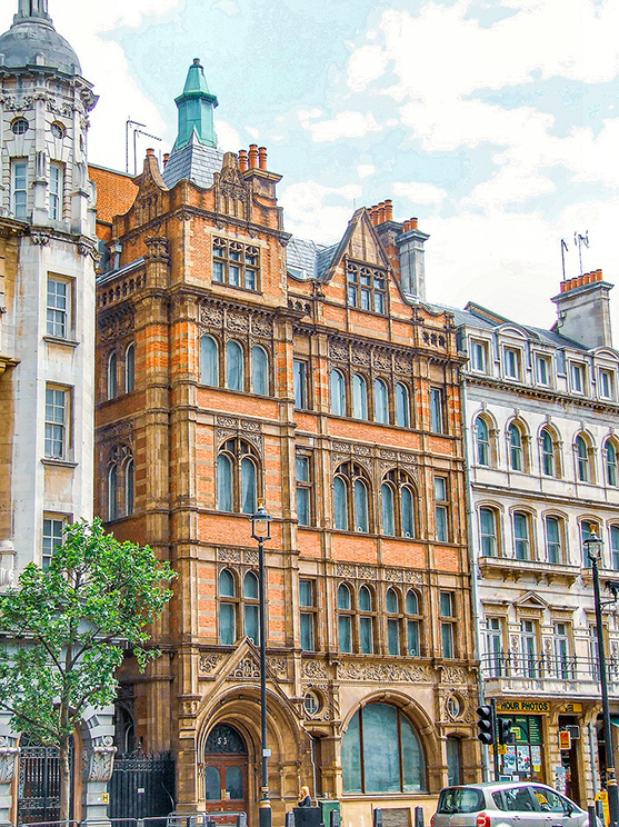 The image depicts a street scene with a prominent, ornate building made of red brick with intricate architectural details. The building features arched windows and decorative stonework. It is situated between other buildings of varying architectural styles. There is a tree and a car parked in front of the building, and the sky is partly cloudy.