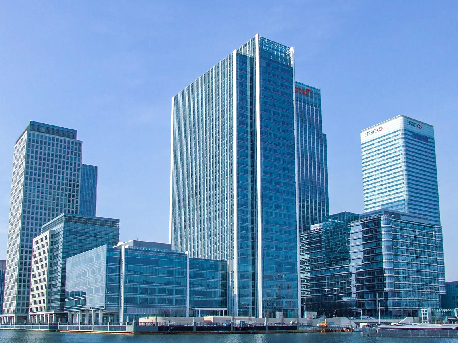 The image depicts a cluster of modern skyscrapers in a financial district. The tallest building in the center has the logo of HSBC, a global bank, prominently displayed near the top. The buildings are made of glass and steel, reflecting a contemporary architectural style. The scene is set against a clear blue sky, and the buildings are situated along a waterfront, indicating a location near a river or harbor. The overall atmosphere suggests a bustling business environment.