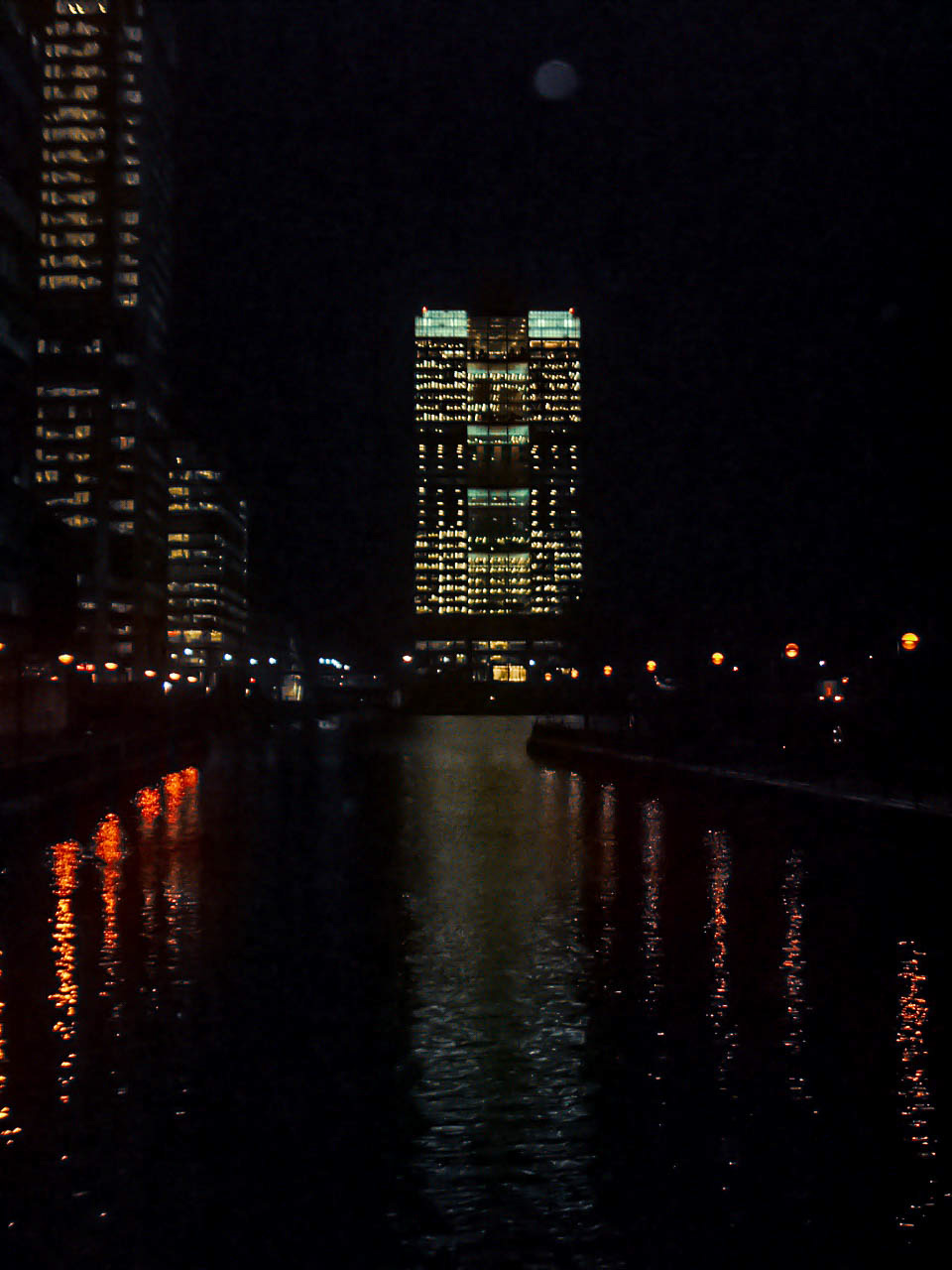 The image depicts a nighttime cityscape with tall buildings illuminated by lights. The reflections of the lights can be seen in the water in the foreground, creating a serene and picturesque scene. The buildings are modern and well-lit, suggesting a bustling urban environment.