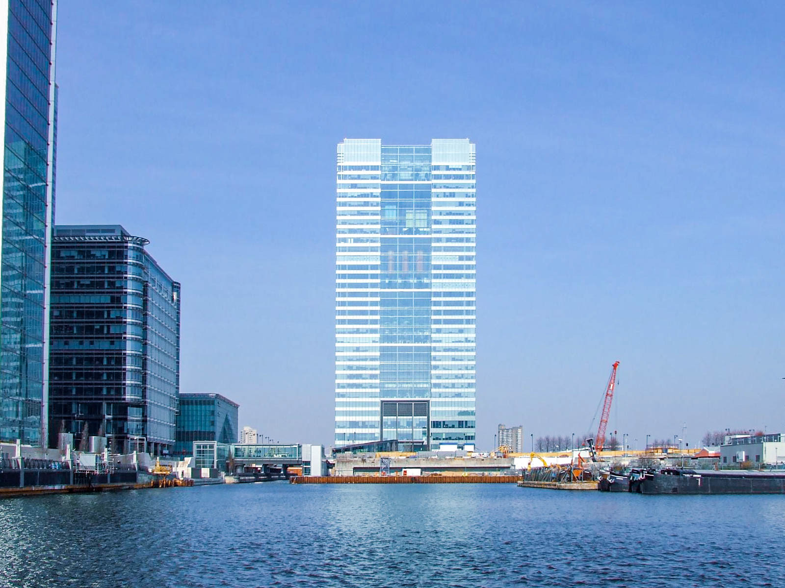 The image depicts a modern urban waterfront scene with several high-rise buildings. The tallest building is centrally located and has a reflective glass facade. To the left, there is another tall building with a mix of glass and darker panels. In the foreground, there is a body of water with some construction activity visible on the right side of the image, including cranes and construction equipment. The sky is clear, indicating a sunny day.