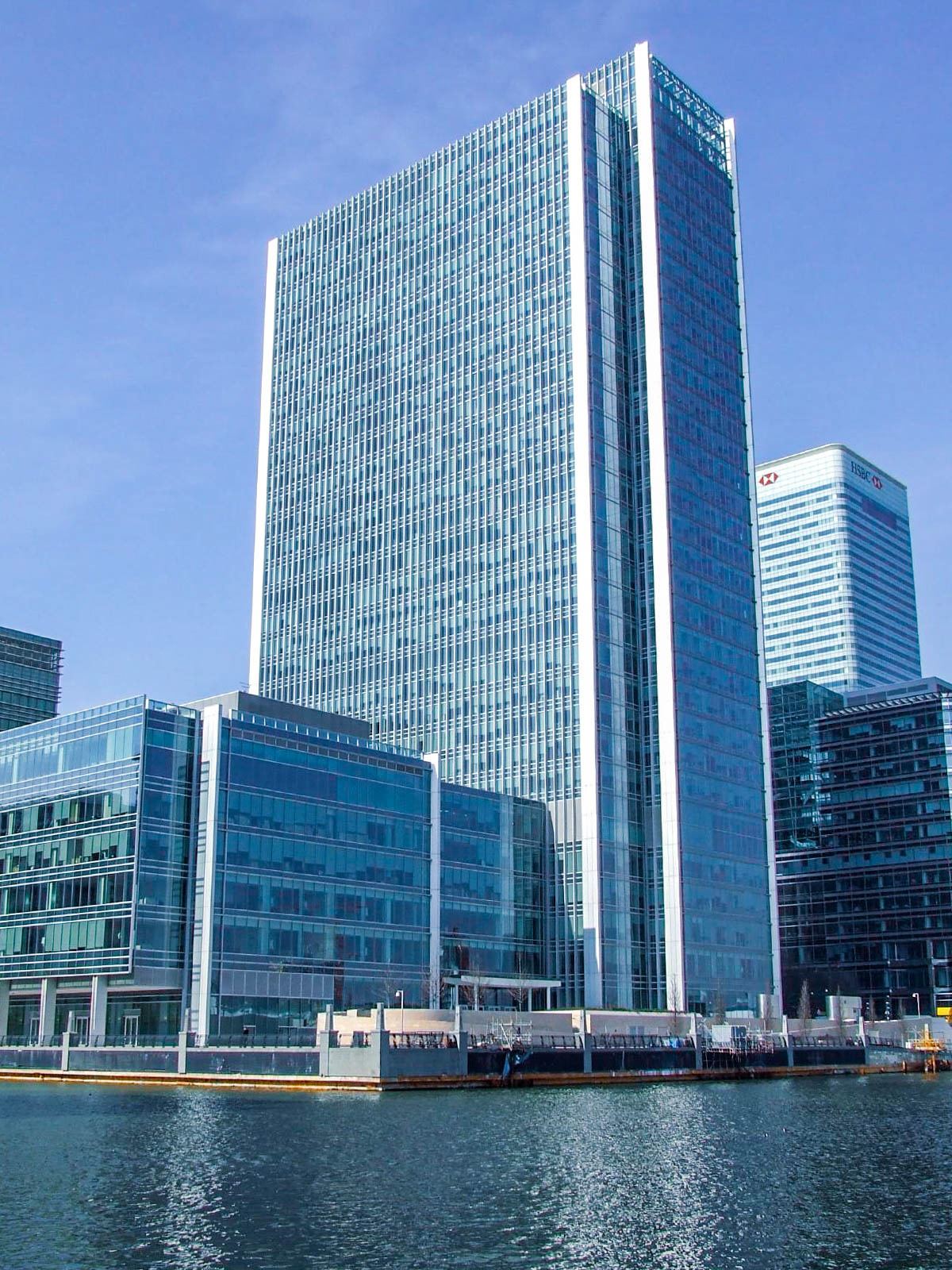 The image depicts a modern urban waterfront scene featuring several high-rise buildings. The central structure is a tall, rectangular skyscraper with a glass facade, reflecting the sky and surrounding buildings. To the right, another prominent building displays the HSBC logo at the top. In the foreground, there are smaller glass buildings adjacent to the water, creating a sleek and contemporary architectural aesthetic. The water in the foreground suggests the buildings are located near a river or harbor.