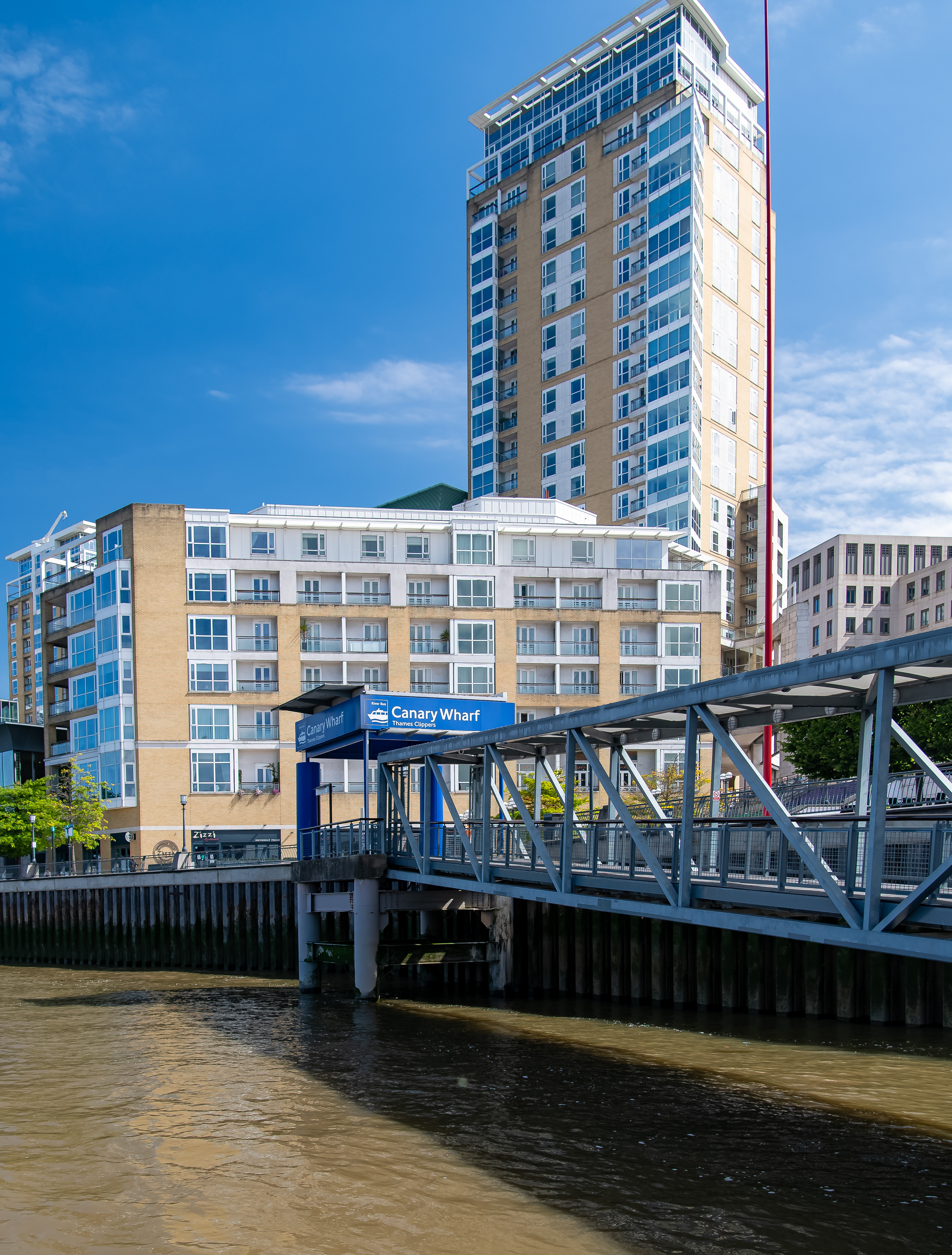The image shows a waterfront scene at Canary Wharf, featuring modern high-rise buildings and a pedestrian bridge leading to a dock. The sky is clear with a few clouds, and the water is calm. The prominent structure is a tall building with a mix of glass and brick facade. The sign indicates a Thames Clippers stop, suggesting transportation services.