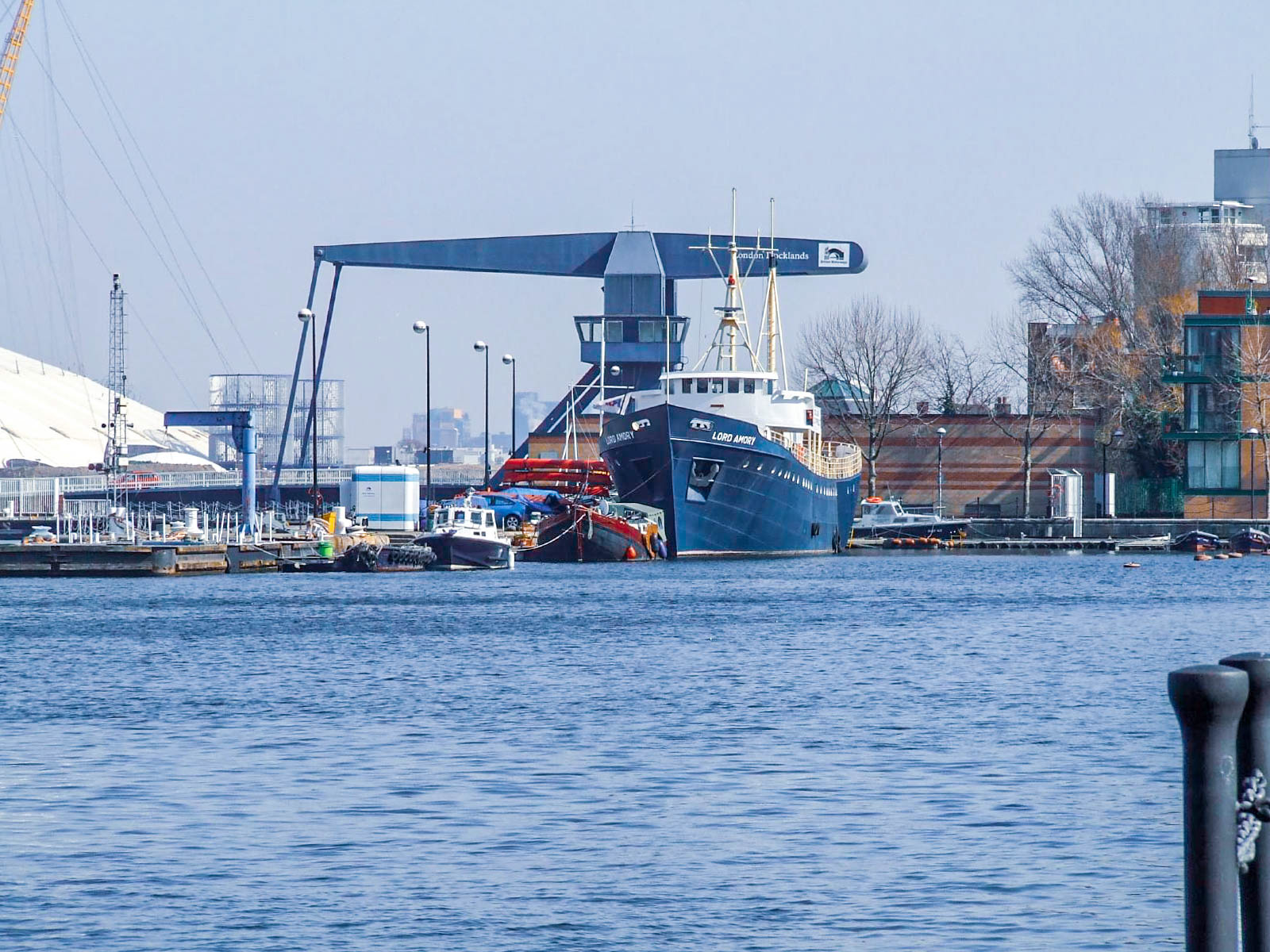 The image depicts a dock scene with a large ship named 'Lord Ashfield' belonging to the company 'London Towlands'. The ship is moored at a pier, with various smaller boats and equipment around it. The background shows industrial buildings and cranes, indicating a working dockyard environment.