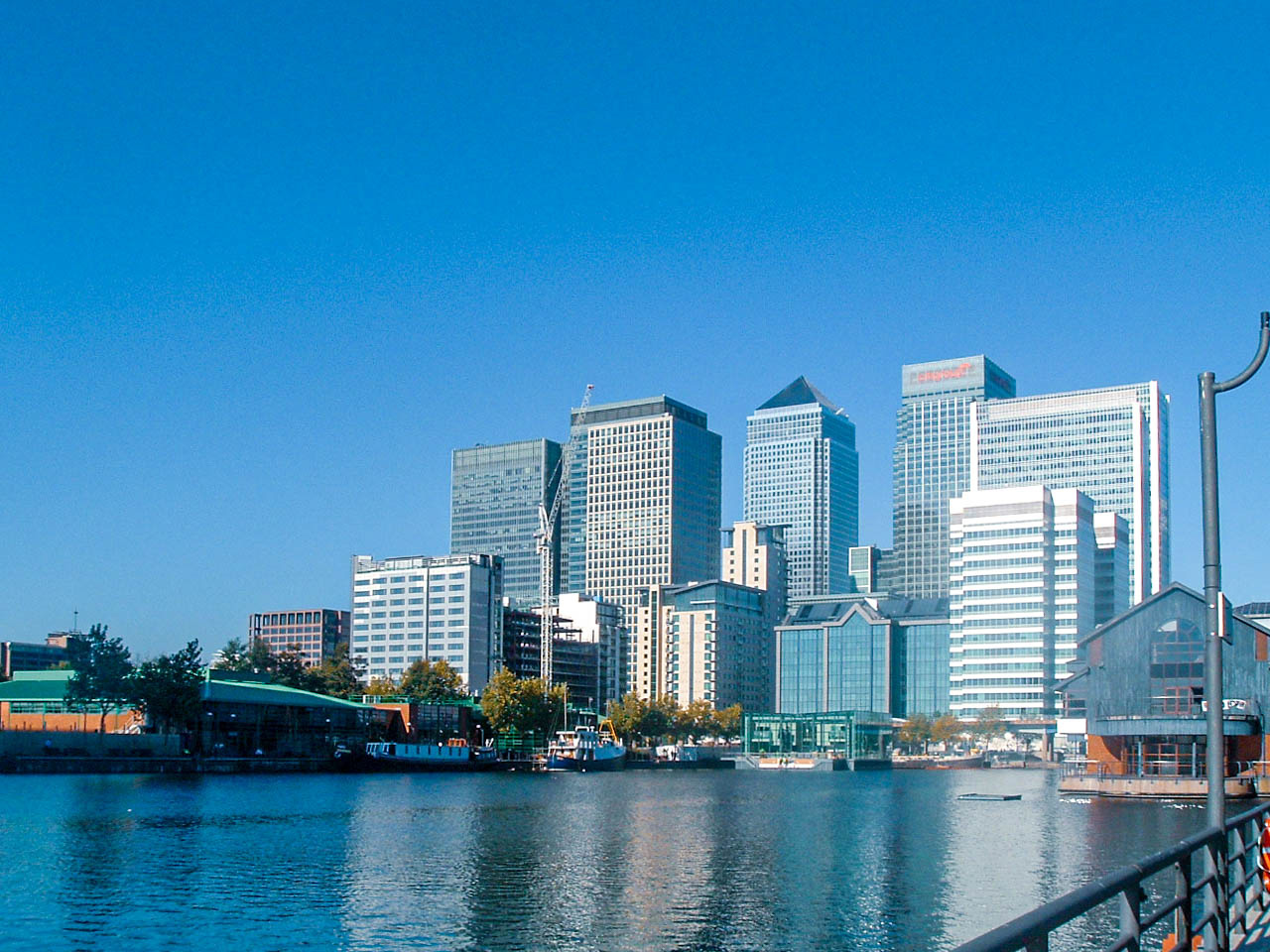 The image depicts a modern cityscape with tall skyscrapers and high-rise buildings along a waterfront. The buildings are primarily made of glass and steel, reflecting a contemporary architectural style. The sky is clear and blue, indicating a sunny day. In the foreground, there is a body of water with some boats and a pier. The scene suggests a bustling urban area, likely a financial or business district.