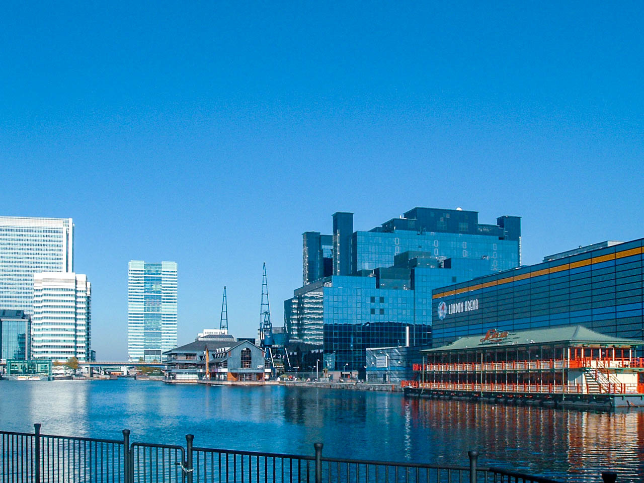 The image depicts a waterfront cityscape with modern buildings and the London Arena prominently displayed. The scene includes a mix of high-rise office buildings, a large glass-fronted structure, and a smaller building with a distinctive roof. The water in the foreground reflects the buildings, and the sky is clear and blue.