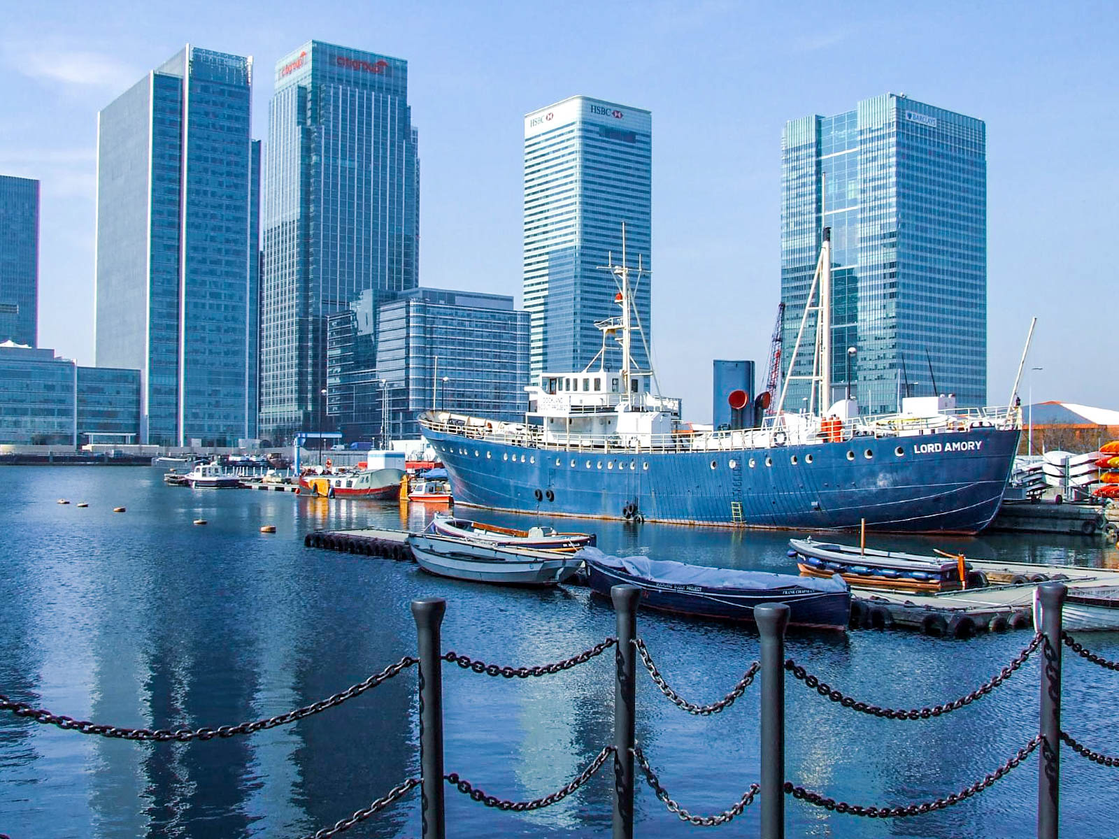 The image depicts a modern cityscape with several high-rise buildings, including those labeled 'HSBC' and 'Nordea'. In the foreground, there is a large, vintage ship named 'Lord Amory' docked at a marina. Smaller boats are also moored nearby, and the scene is framed by a chain-linked barrier in the foreground. The overall atmosphere suggests a blend of historical and contemporary elements, likely in a financial or business district near a harbor.